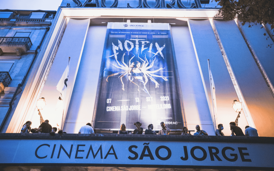 Night view of Cinema São Jorge in Lisbon during MOTELX, the Lisbon International Horror Film Festival. A large poster of the festival's 16th edition hangs above the entrance, depicting a surreal multi-limbed figure with dramatic lighting. People are gathered on the balcony, contributing to the lively cultural atmosphere of the event.