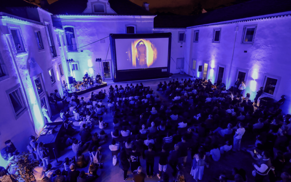 Outdoor horror film screening during MOTELX, the Lisbon International Horror Film Festival. A large audience is seated in a courtyard illuminated by atmospheric blue lighting, watching a scene from a horror movie on a giant screen. Live musicians perform beside the screen, enhancing the immersive experience.