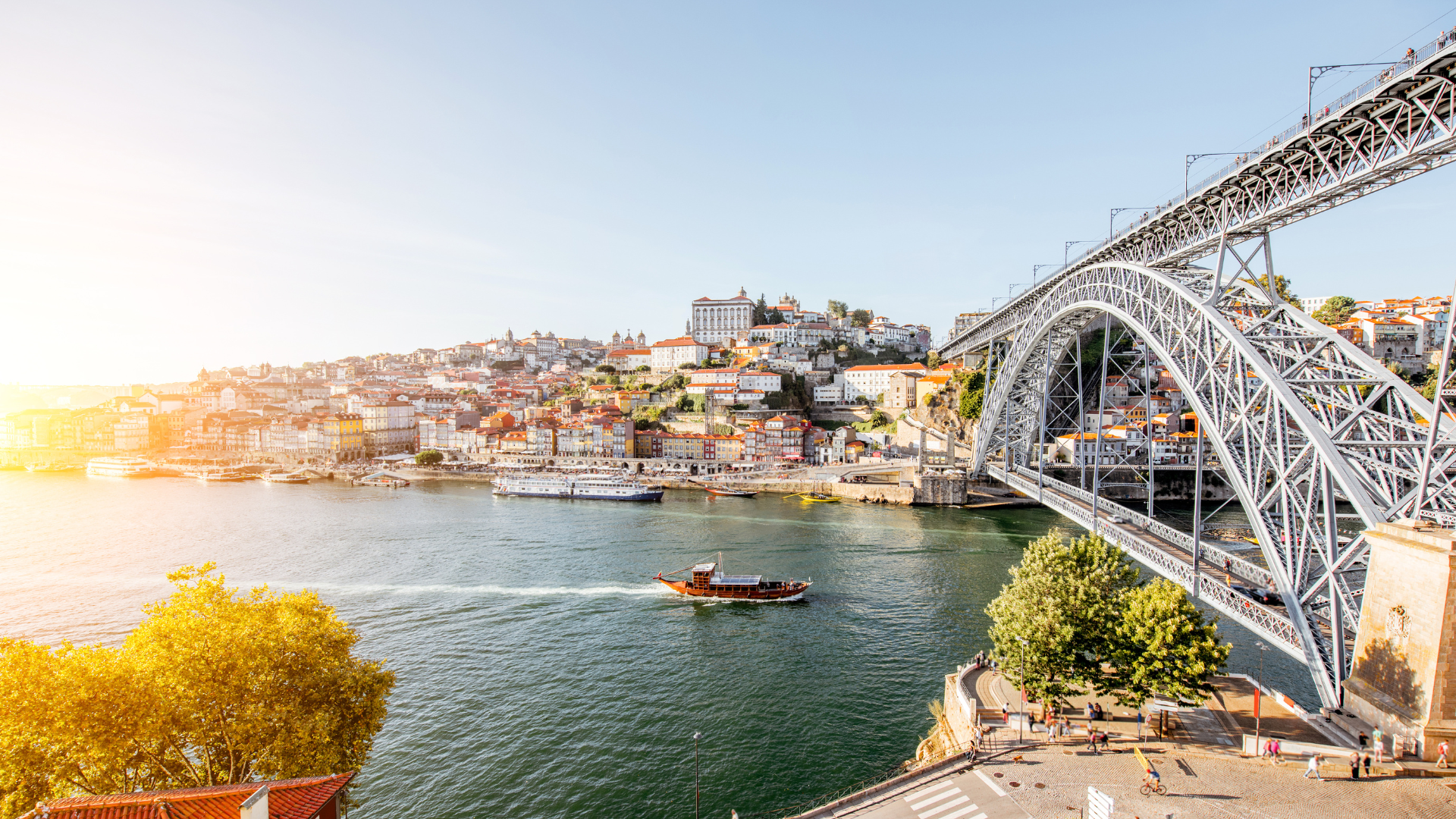 Panoramic view of Luís I Bridge and Porto’s Ribeira district at sunset, with boats sailing on the Douro River.