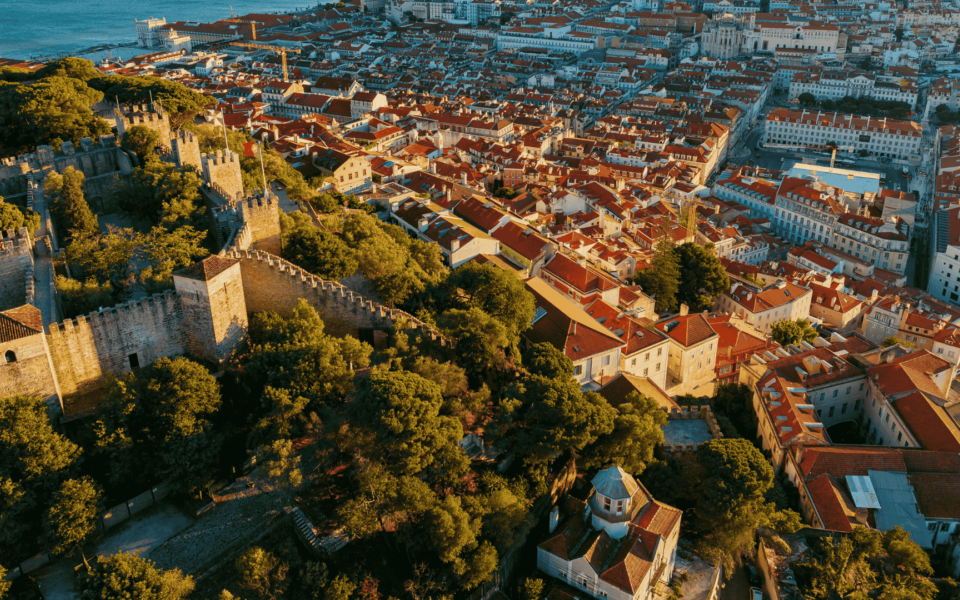 Aerial panoramic view of São Jorge Castle in Lisbon, Portugal, with its medieval walls surrounded by trees and overlooking the historic city center and rooftops.