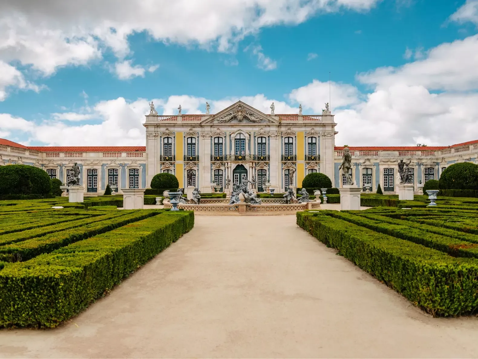 Baroque façade of the National Palace of Queluz with manicured gardens and statues under a blue sky with clouds.