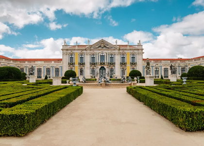 Baroque façade of the National Palace of Queluz with manicured gardens and statues under a blue sky with clouds.