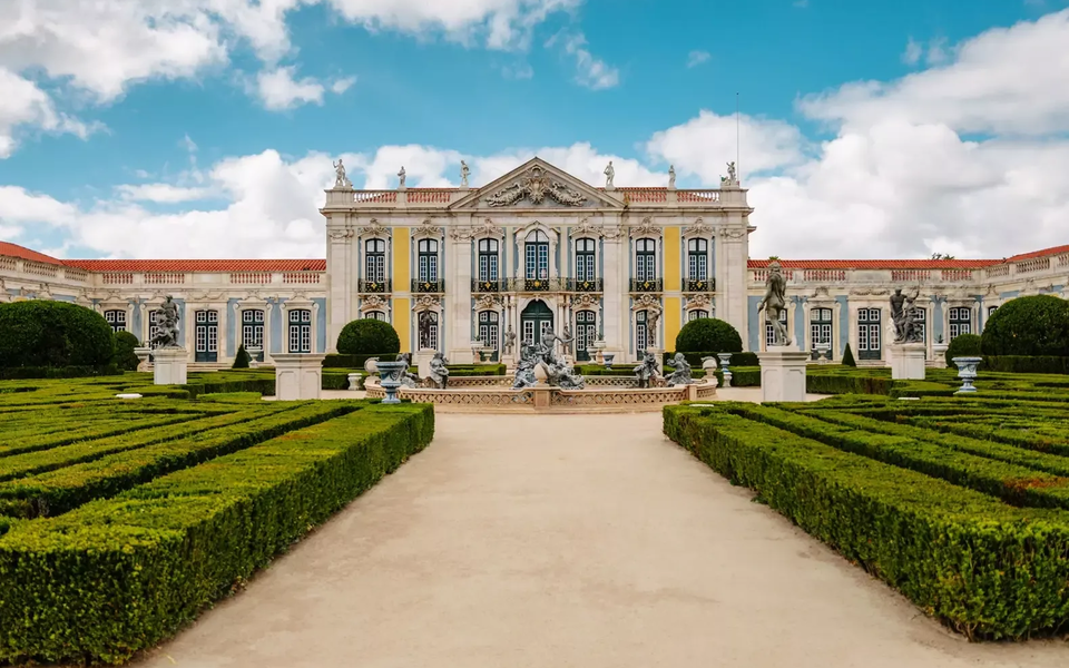 Baroque façade of the National Palace of Queluz with manicured gardens and statues under a blue sky with clouds.