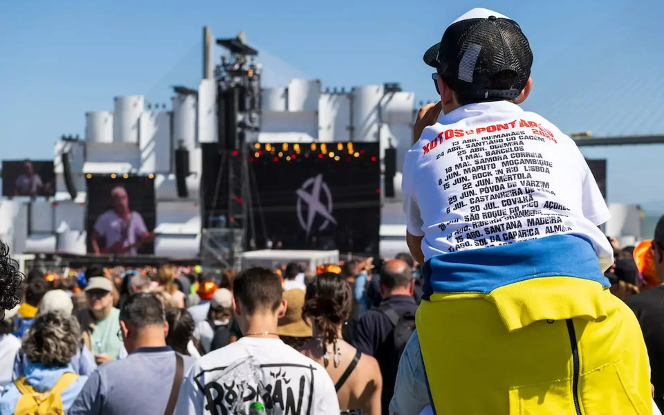A child wearing a tour dates shirt sits on an adult’s shoulders watching a daytime concert at Rock in Rio Lisboa, with the main stage and crowd in view