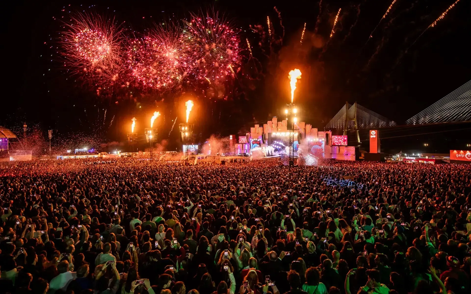 Massive crowd at Rock in Rio Lisboa enjoying a night concert with fireworks, flames, and the Vasco da Gama bridge in the background