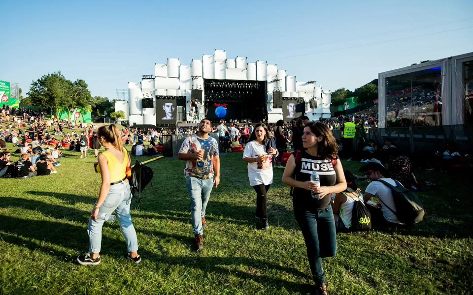 People relaxing on the grass at Rock in Rio Lisboa during daylight, with the main stage in the background and a casual festival vibe