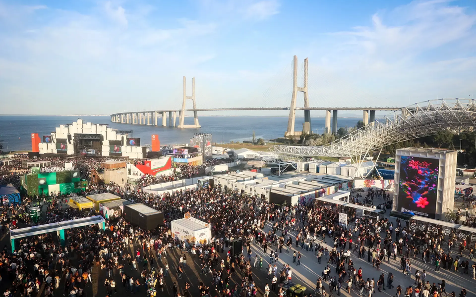 Aerial view of Rock in Rio Lisboa festival grounds with the stage, food stalls, and the Vasco da Gama bridge over the Tagus River in the background