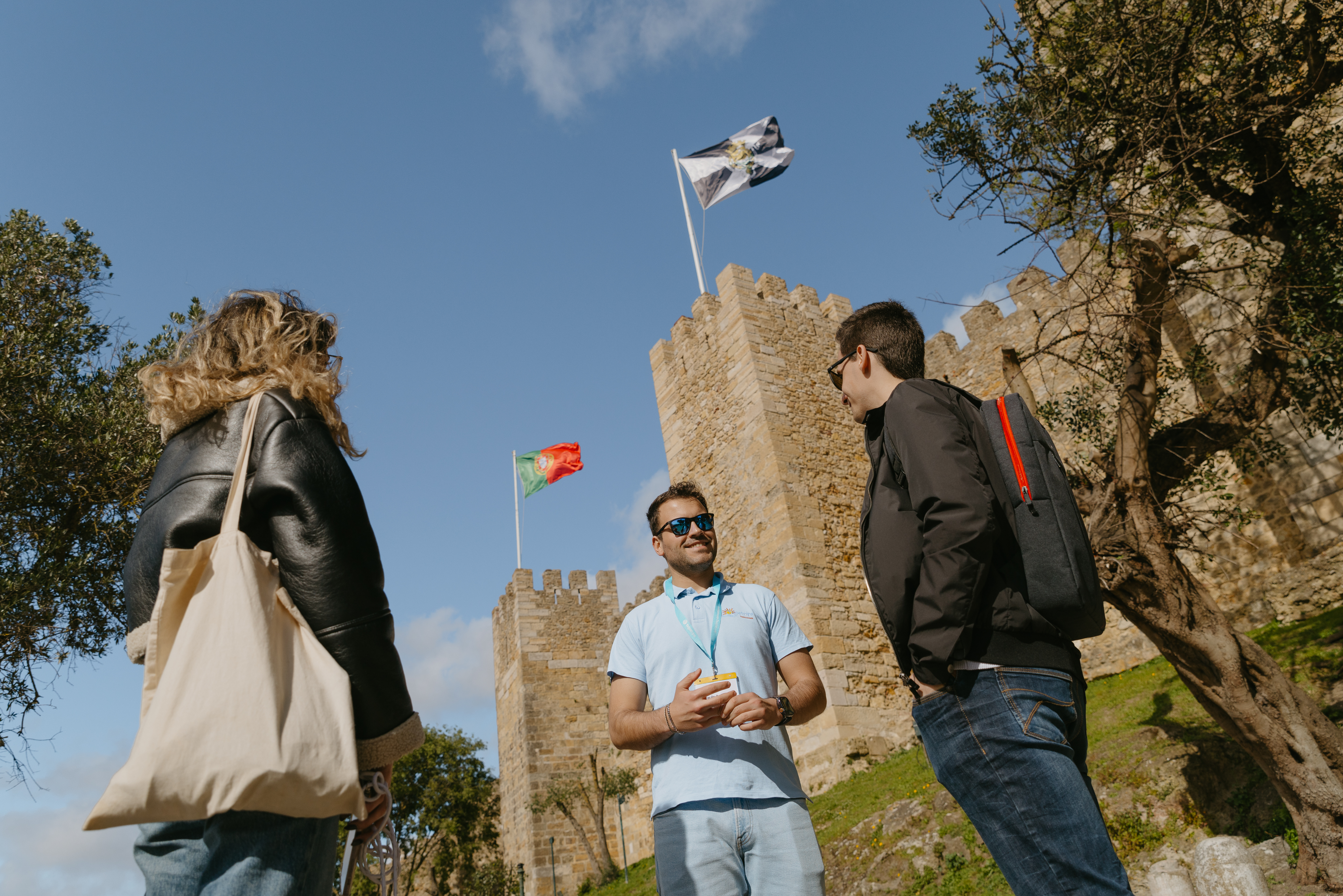 A guide in blue stands with two visitors in front of the majestic Castelo de São Jorge, showcasing its grandeur.