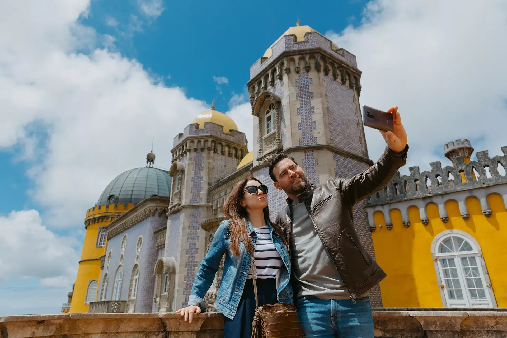 A happy couple takes a selfie in front of the stunning Pena Palace in Sintra, Portugal, capturing a beautiful moment together.