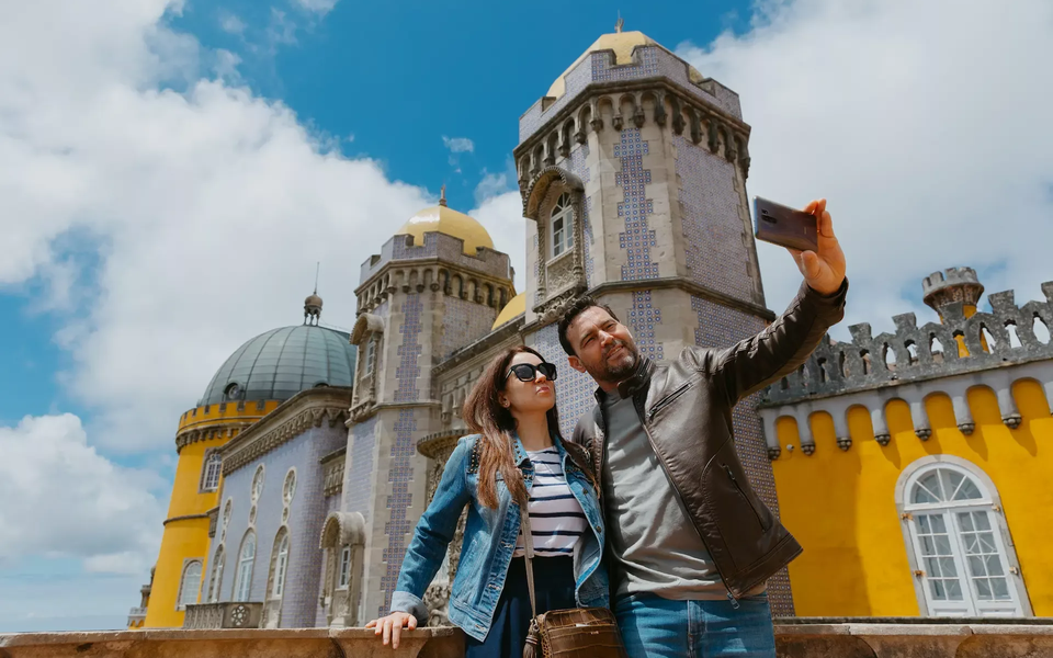 A happy couple takes a selfie in front of the stunning Pena Palace in Sintra, Portugal, capturing a beautiful moment together.