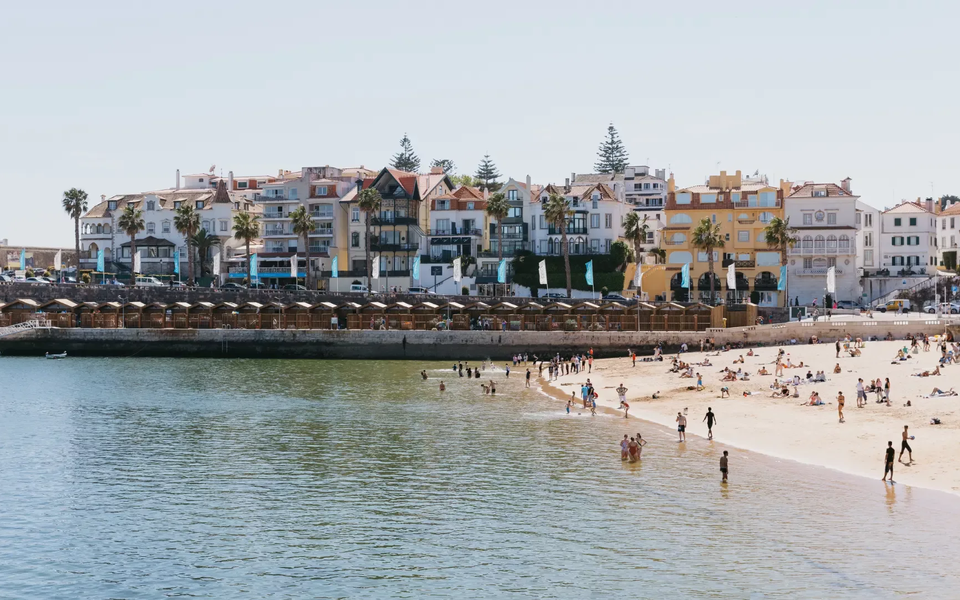 People enjoying the sunny beach in Cascais with colorful houses and palm trees in the background.