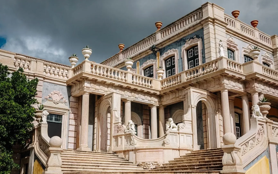 Facade of the National Palace of Queluz with ornate baroque architecture and dramatic staircase under a cloudy sky.