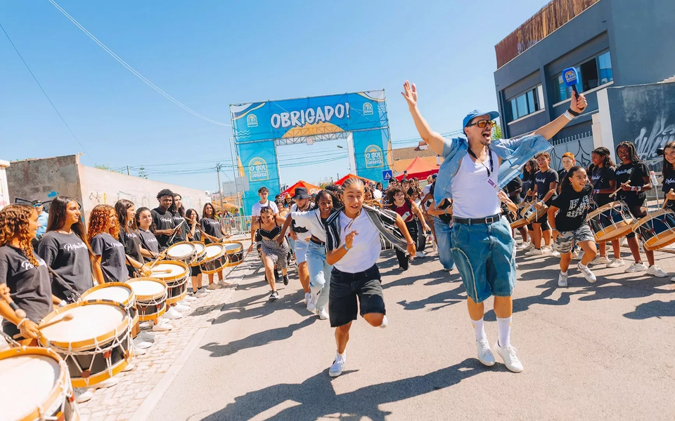People running with joy at the entrance of O Sol da Caparica Festival, with drummers performing under the summer sun in Portugal.