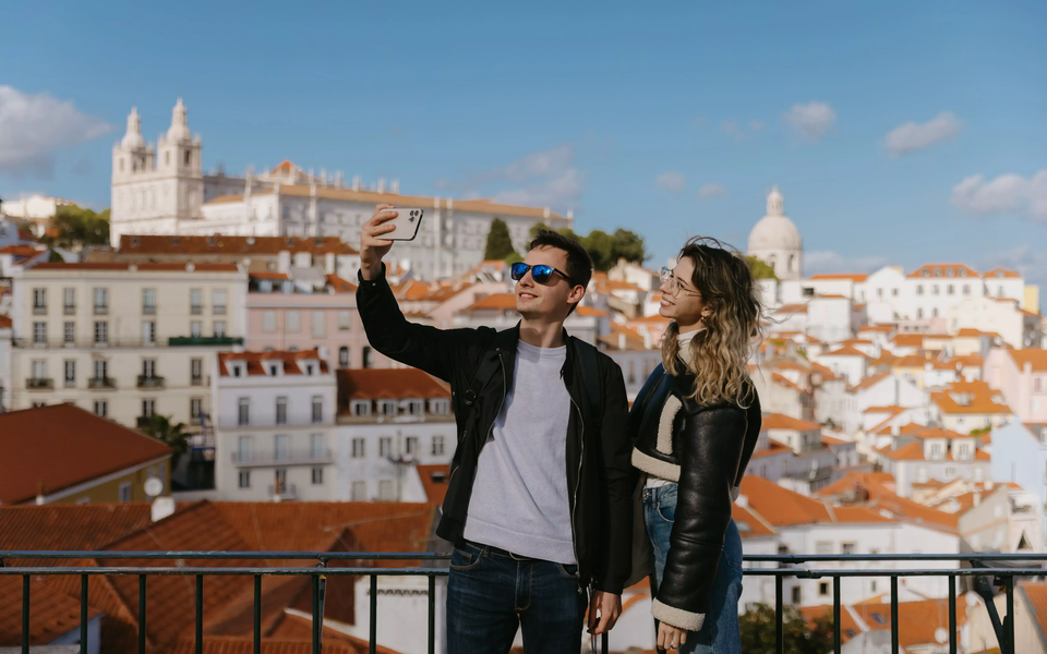 Young couple taking a selfie at Portas do Sol viewpoint with a scenic view of Alfama rooftops, the National Pantheon and the Church of São Vicente de Fora in the background.
