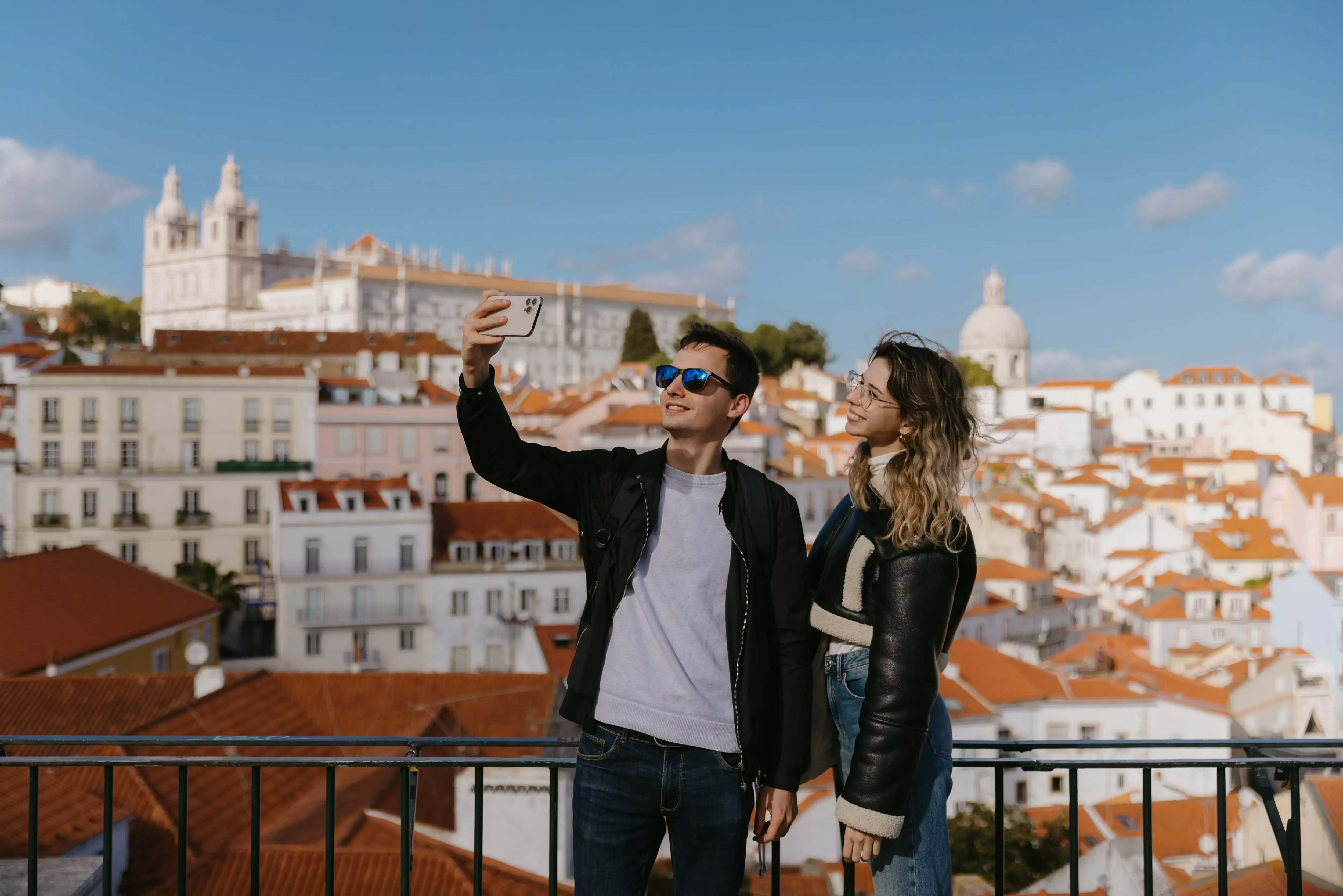 Young couple taking a selfie at Portas do Sol viewpoint with a scenic view of Alfama rooftops, the National Pantheon and the Church of São Vicente de Fora in the background.