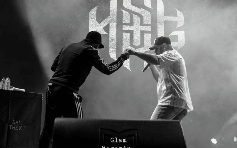 Black and white photo of Portuguese rapper Sam the Kid performing live on stage, sharing a handshake with another artist during a powerful hip hop set. The iconic STK logo is illuminated in the background.
