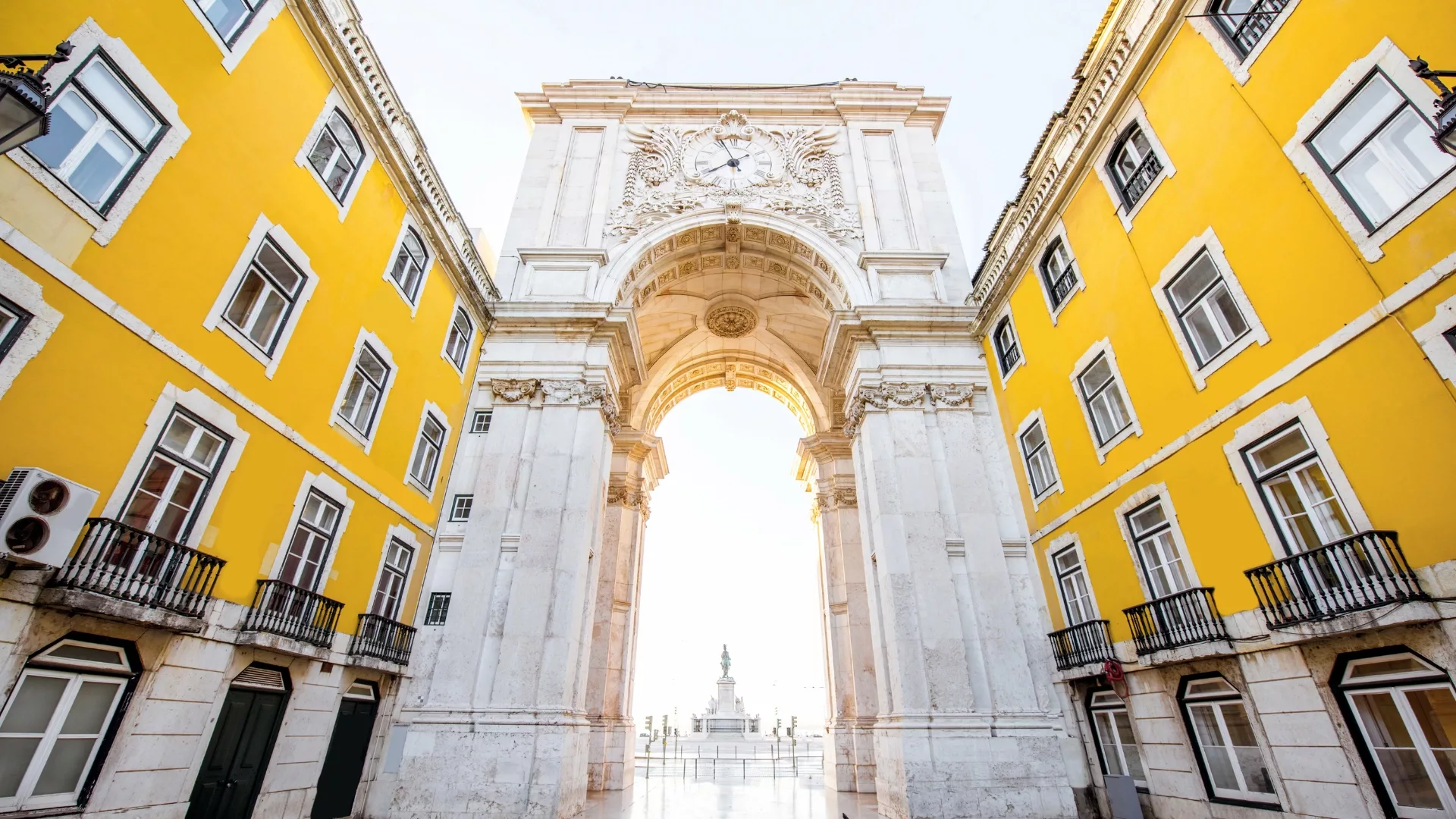 View of the Rua Augusta Arch in Lisbon, framed by yellow Pombaline buildings, with the statue of King José I in the background.