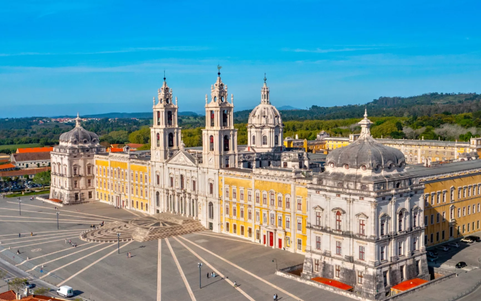 Aerial view of the Mafra National Palace in Portugal, showcasing its Baroque architecture, twin bell towers, and expansive courtyard surrounded by green landscape.