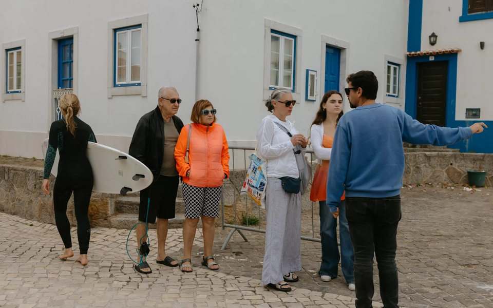 Group of tourists and a barefoot surfer in a picturesque street of Ericeira, Portugal, surrounded by whitewashed houses with blue accents.