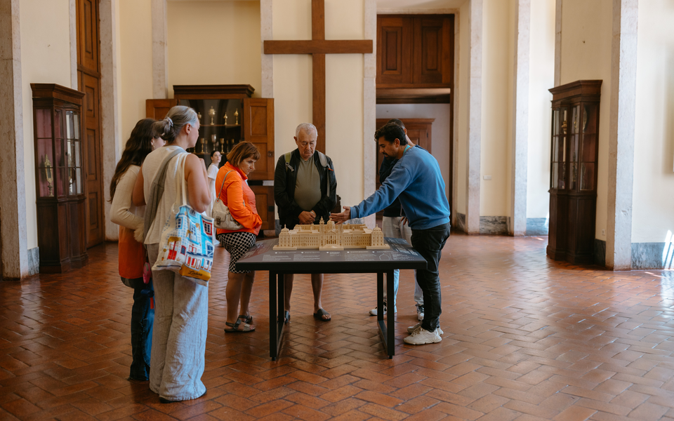 Guide explaining the architectural model of Mafra National Palace to a group of visitors inside one of its historic rooms.