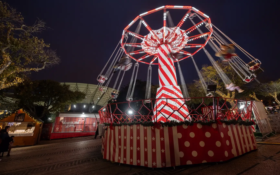 Nighttime view of a spinning carousel swing ride at Wonderland Lisboa, decorated with red and white candy stripes and surrounded by holiday lights and stalls.